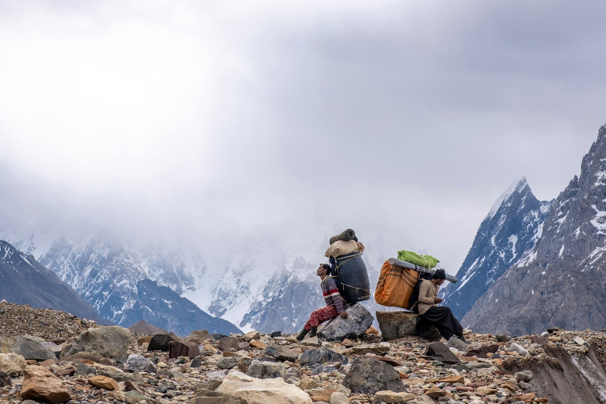 Trekker with a backpack on the rocky moraine of the Baltoro Glacier, looking toward a snow-covered Karakoram peak