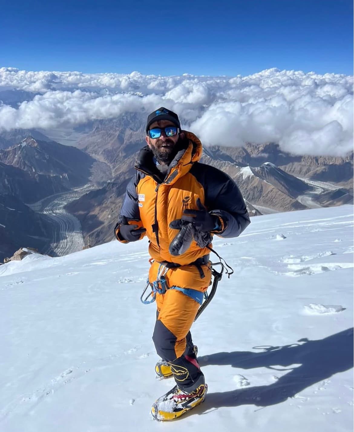 Sohail Sakhi standing in an orange down suit on a snow summit above a sea of cloud
